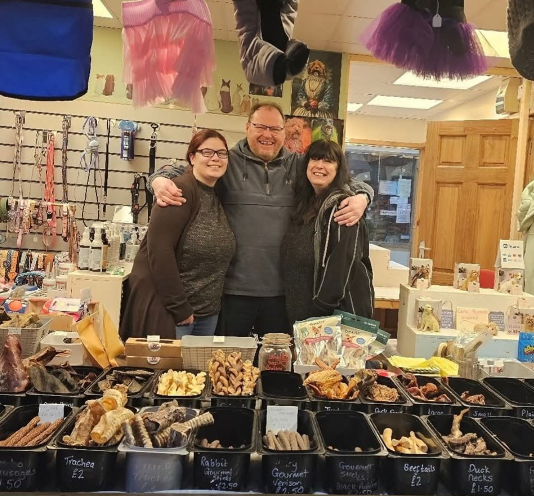 A man stands in the middle of a dog treat shop with his arms around two women, one his wofe one his daighter. They are smiling.